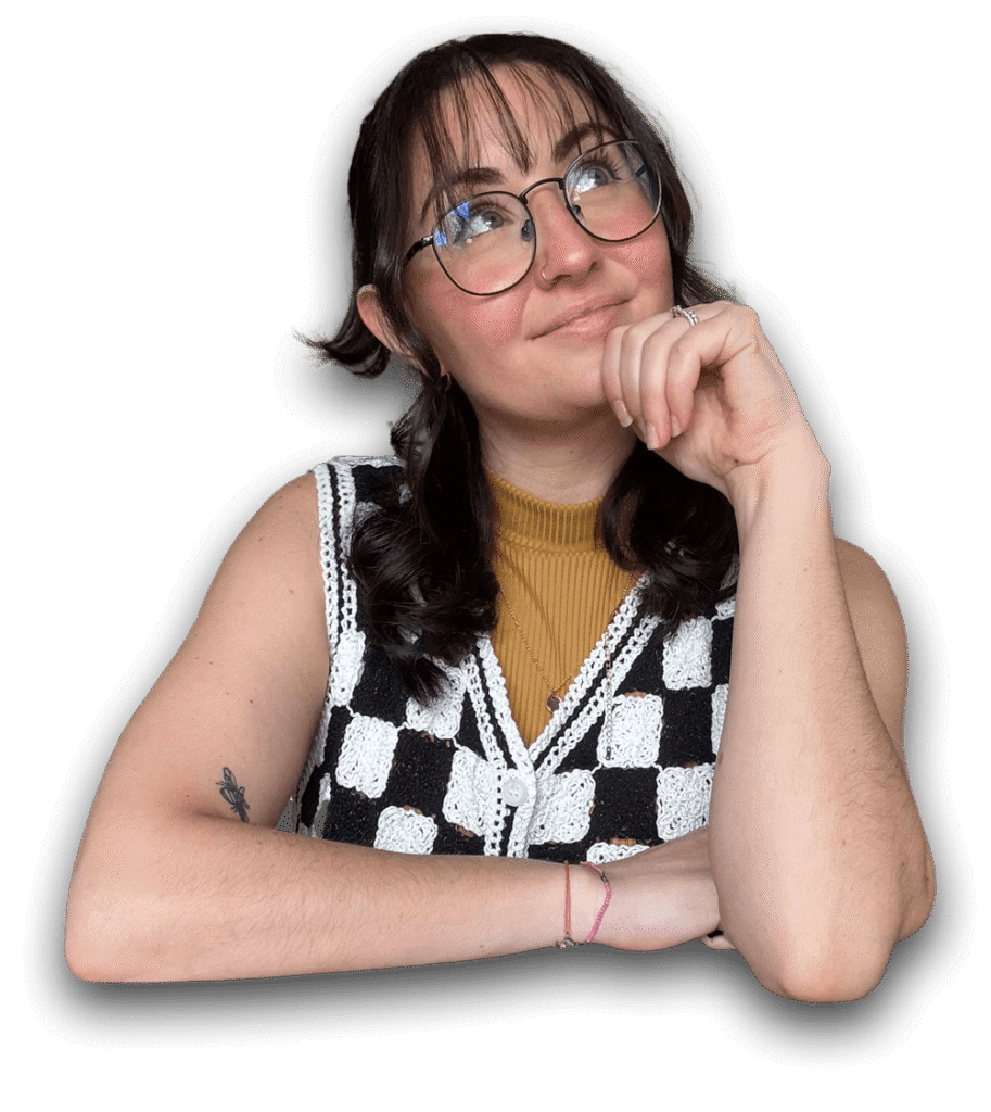A person with glasses and wavy brown hair is smiling thoughtfully. They wear a black and white checker vest over a mustard shirt against a black backdrop.